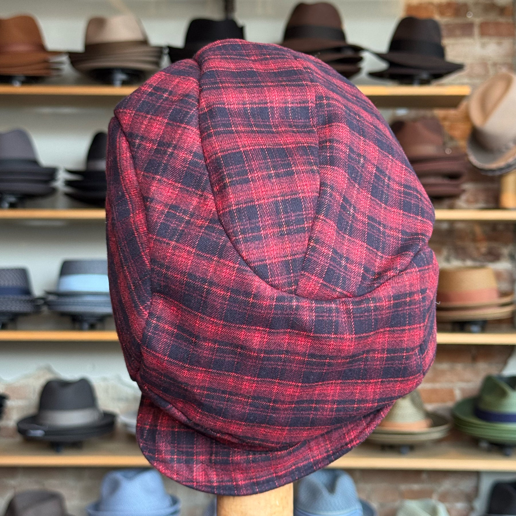Plaid Funk Master Cap in a red and black plaid pattern displayed on a stand with shelves of various hats in the background.