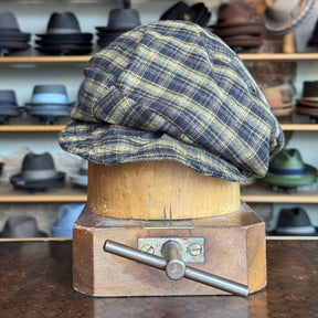 Plaid Funk Master Cap in muted yellow and navy plaid pattern displayed on a wooden hat block with various hats in the background.