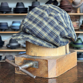 Plaid Funk Master Cap in yellow and navy plaid pattern displayed on a wooden hat block, with various hats blurred in the background, showcasing a classic newsboy style.