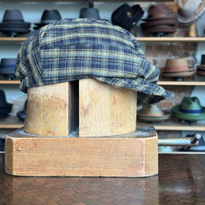 Plaid Funk Master Cap in dark blue and yellow plaid pattern displayed on a wooden hat block with various hats blurred in the background.