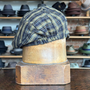 Plaid Funk Master Cap with a classic plaid pattern in muted yellow and blue tones, styled as a newsboy cap displayed on a wooden hat block with shelves of various hats in the background.