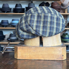 Plaid Funk Master Cap in blue and yellow plaid pattern displayed on a wooden hat block, surrounded by various hats on shelves in the background.