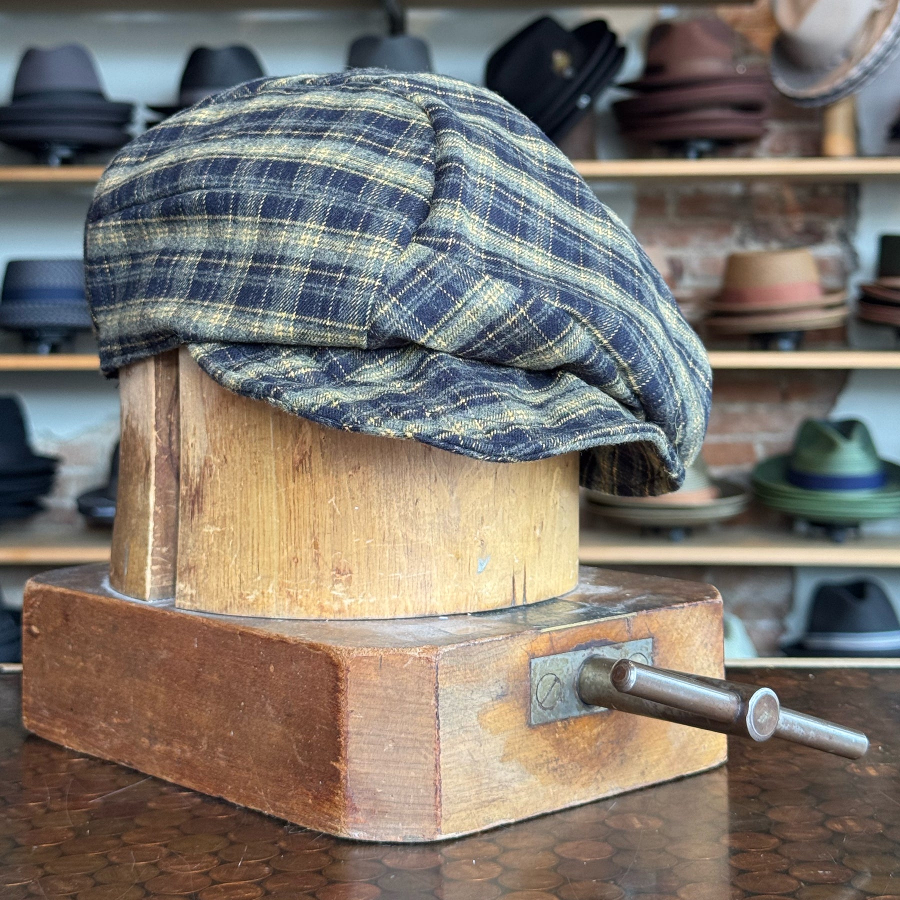 Plaid Funk Master Cap in muted blue and yellow tones displayed on a vintage wooden hat block with hats in the background.