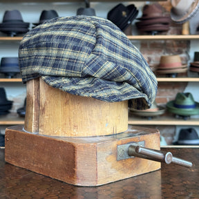 Plaid Funk Master Cap in muted blue and yellow tones displayed on a vintage wooden hat block with hats in the background.