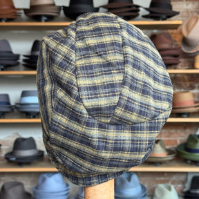 Plaid Funk Master Cap in a muted yellow and gray plaid pattern displayed on a wooden hat stand with a background of various hats on shelves.