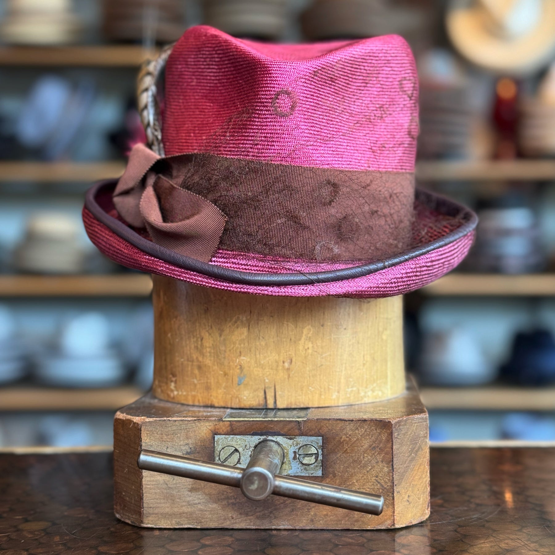 Raspberry Dandy Fedora featuring a textured raspberry pink crown, brown band with a decorative bow, and a feather accent, displayed on a wooden hat block in a hat shop.