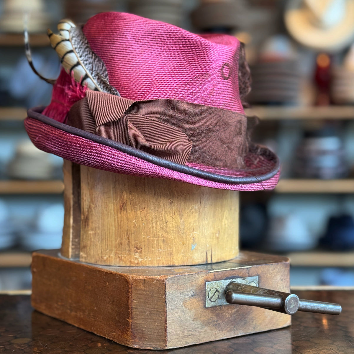 Raspberry Dandy Fedora with a vibrant raspberry color, decorated with a brown textured band and stylish feathers, displayed on a wooden hat block in a cozy hat shop.