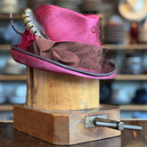 Raspberry Dandy Fedora with a vibrant raspberry color, decorated with a brown textured band and stylish feathers, displayed on a wooden hat block in a cozy hat shop.