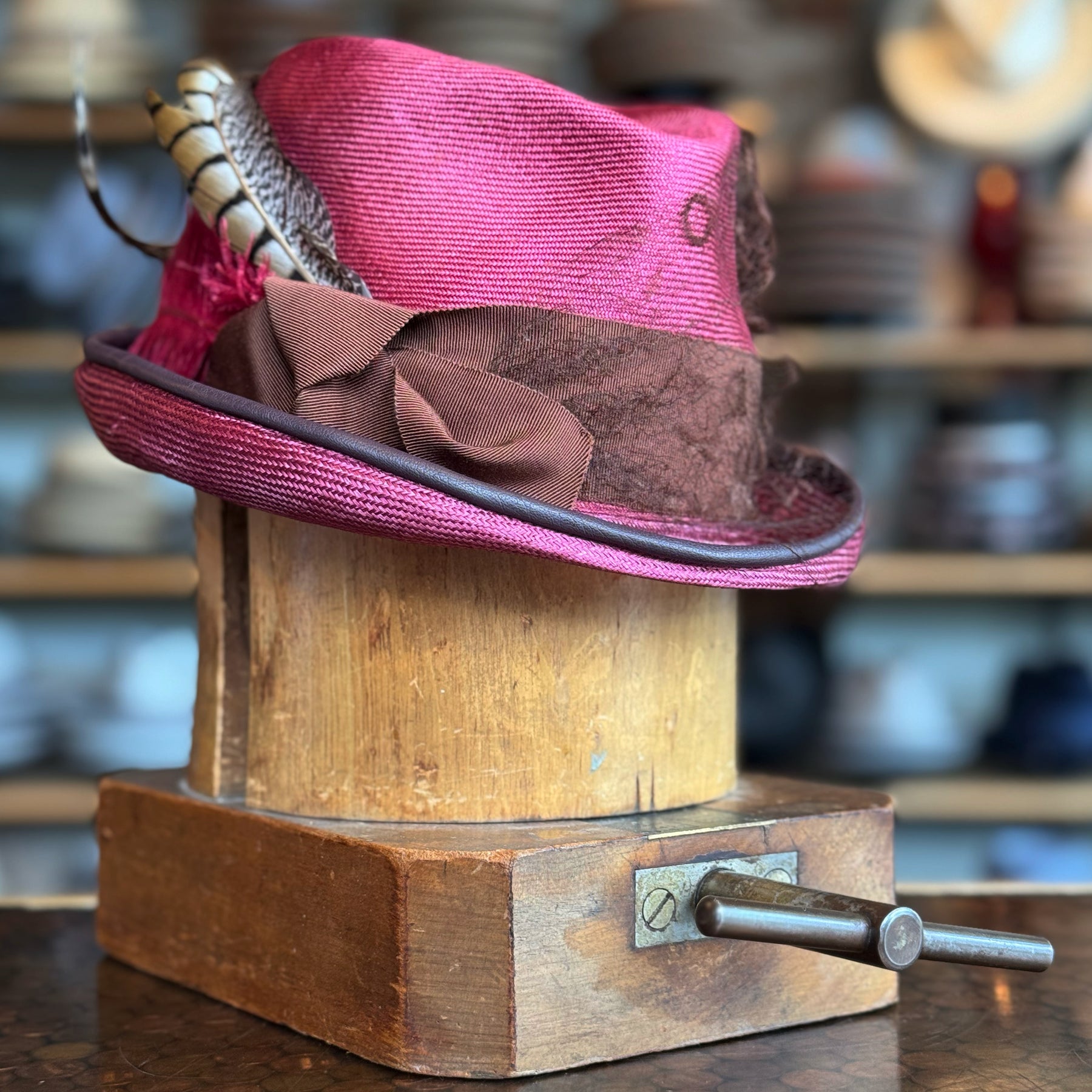 Raspberry Dandy Fedora with a vibrant raspberry color, decorated with a brown textured band and stylish feathers, displayed on a wooden hat block in a cozy hat shop.