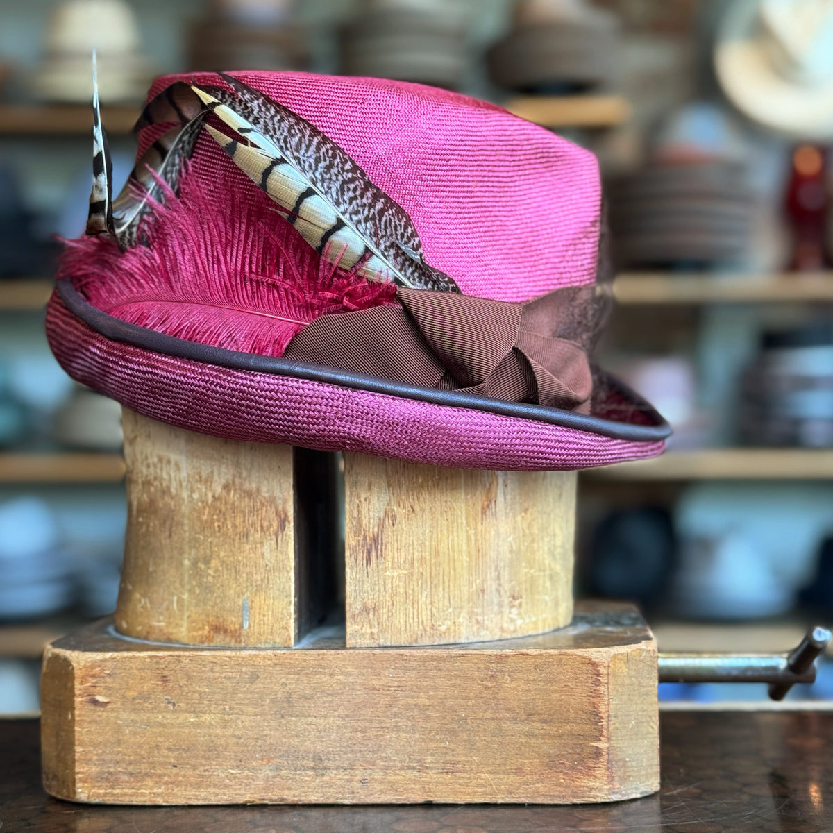 Raspberry Dandy Fedora with a rich raspberry color, decorated with a brown band and adorned with elegant feathers, displayed on a wooden hat block in a hat shop.