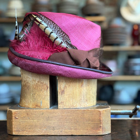 Raspberry Dandy Fedora with a rich raspberry color, decorated with a brown band and adorned with elegant feathers, displayed on a wooden hat block in a hat shop.