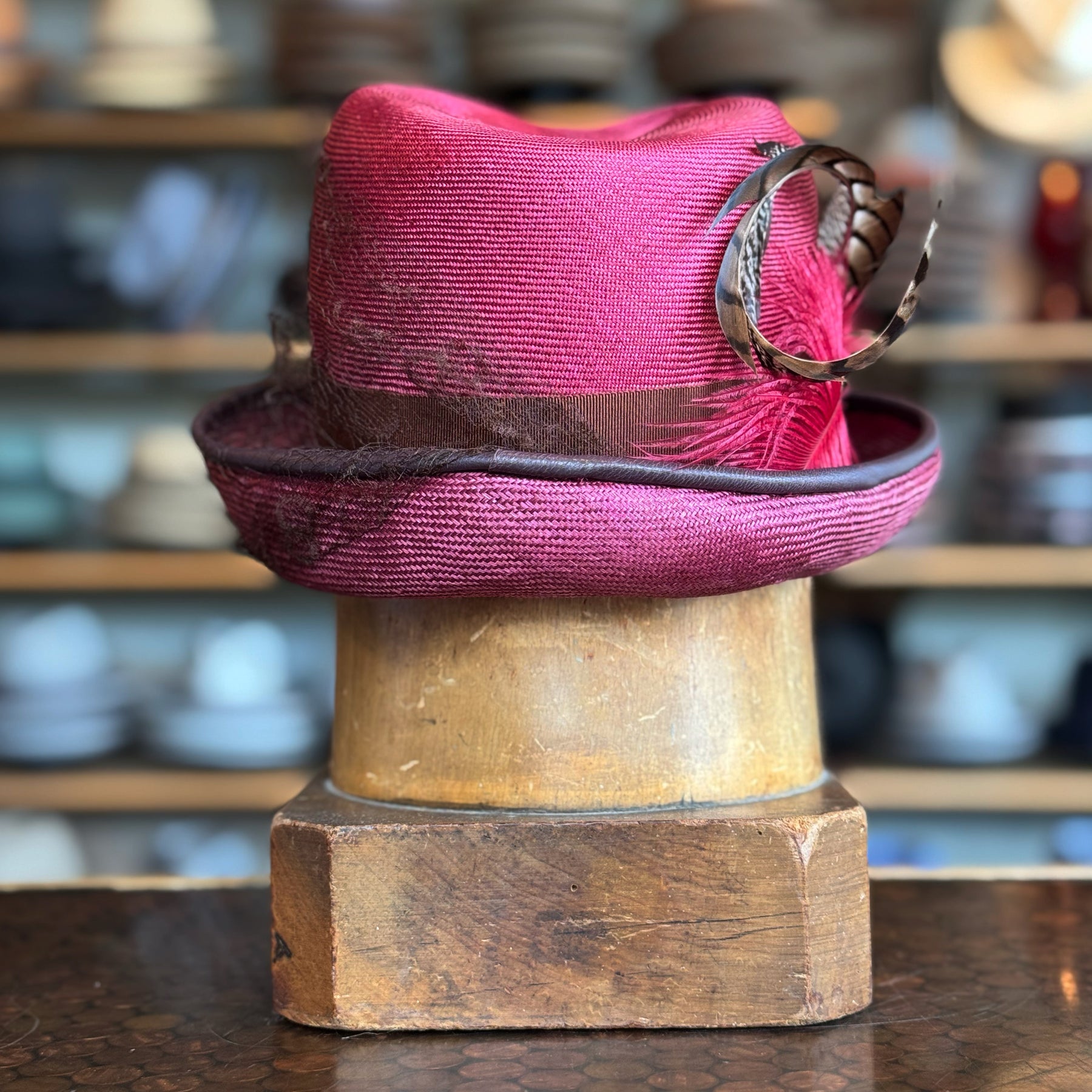 Raspberry Dandy Fedora in vibrant raspberry color with a brown feathered band and decorative feather accent, displayed on a wooden hat stand in a vintage setting.