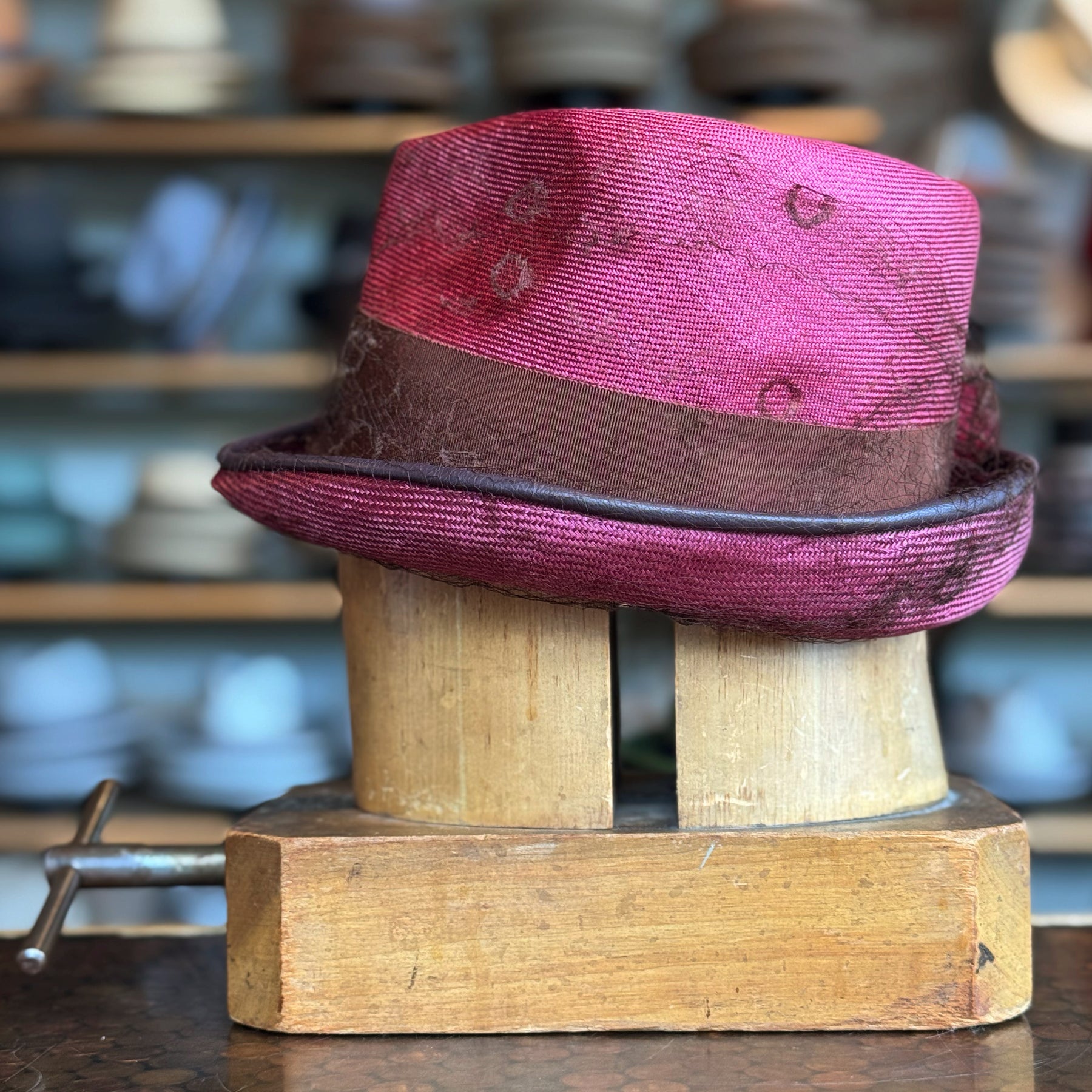 Raspberry Dandy Fedora in a vibrant raspberry color with a brown band, displayed on a wooden hat block with other hats blurred in the background.
