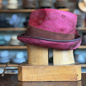 Raspberry Dandy Fedora in a vibrant raspberry color with a brown band, displayed on a wooden hat block with other hats blurred in the background.