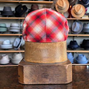 Back view of a Red Check 2-Panel Ivy Cap displayed on a wooden hat block with shelves of various hats in the background