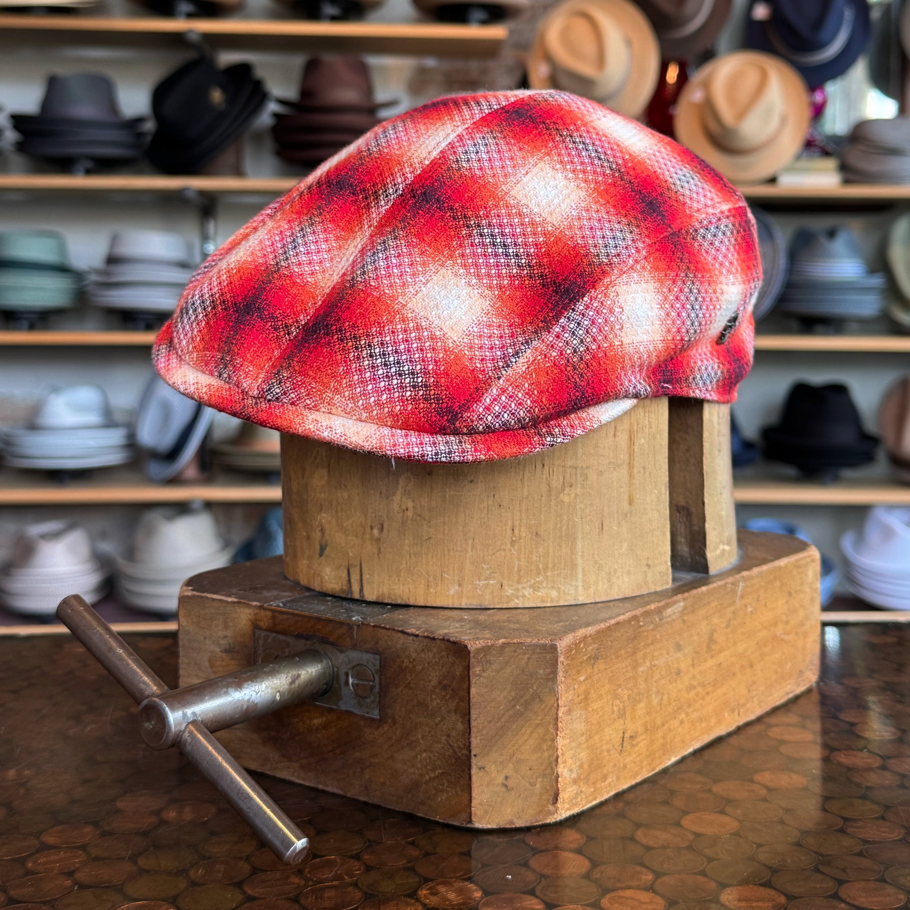 Red Check 2-Panel Ivy Cap displayed on a wooden hat block with a background of various hats, showcasing a vibrant red plaid pattern in a classic flat cap style.