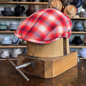 Red Check 2-Panel Ivy Cap displayed on a wooden hat block with a background of various hats, showcasing a vibrant red plaid pattern in a classic flat cap style.