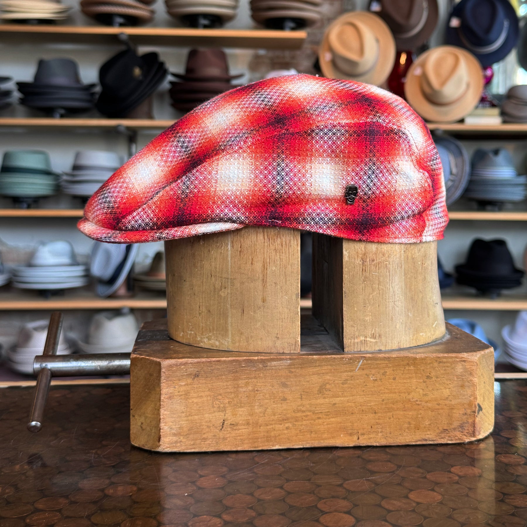 Red Check 2-Panel Ivy Cap with a vibrant red and white check pattern displayed on a wooden hat stand in a store filled with various styles of caps and hats in the background.