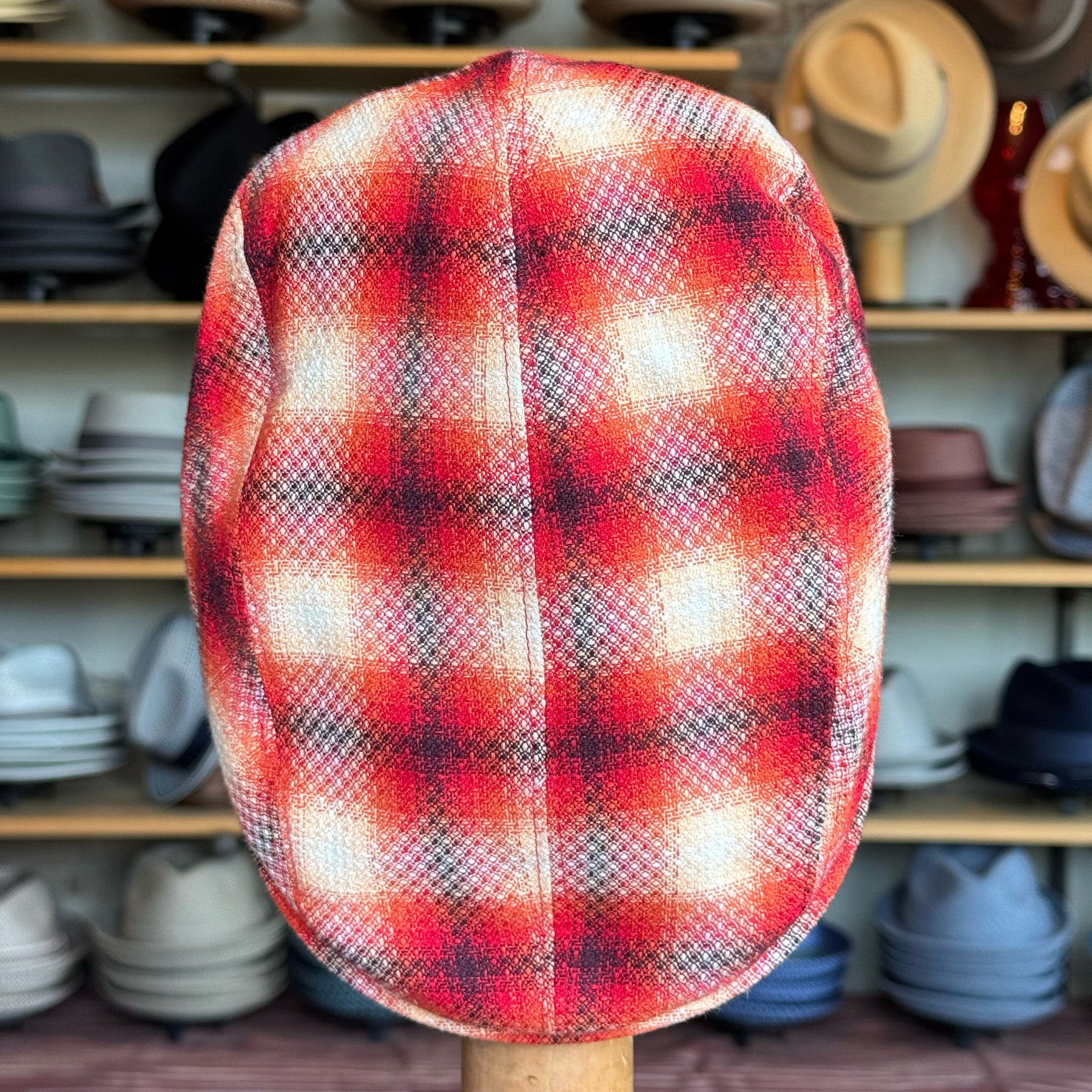 Red Check 2-Panel Ivy Cap showcasing a vibrant red plaid pattern with subtle black and white accents, displayed on a stand in front of shelves filled with various brim hats and caps.