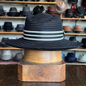 Sabine Fedora in black with a wide brim and white striped band, displayed on a wooden hat block with shelves of various hats in the background.