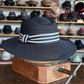 Sabine Fedora in black woven material with a white and black striped band, displayed on a wooden hat block in a hat shop filled with various hats in the background.