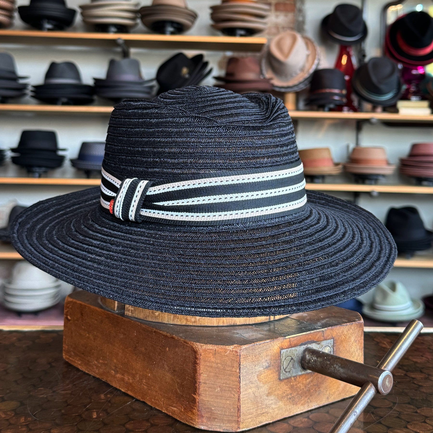 Sabine Fedora in black woven material with a white and black striped band, displayed on a wooden hat block in a hat shop filled with various hats in the background.