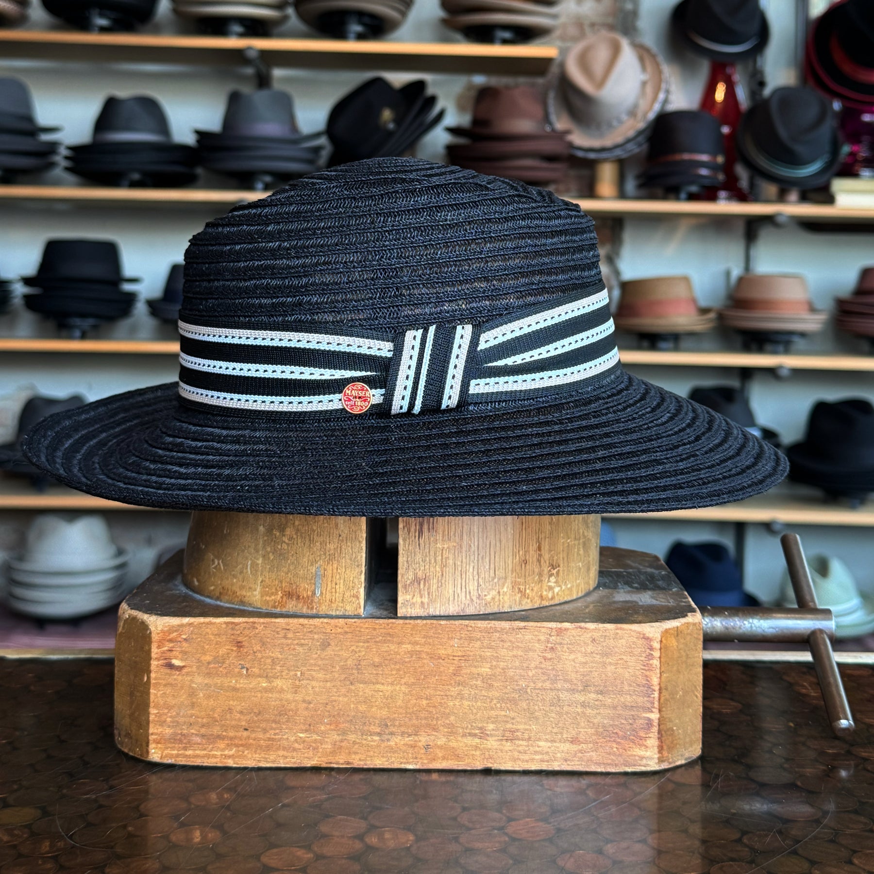 Black woven fedora hat with a striped black and white ribbon band, displayed on a wooden block with shelves of various hats in the background.