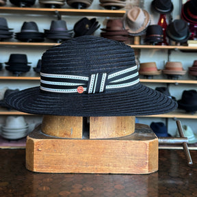 Black woven fedora hat with a striped black and white ribbon band, displayed on a wooden block with shelves of various hats in the background.