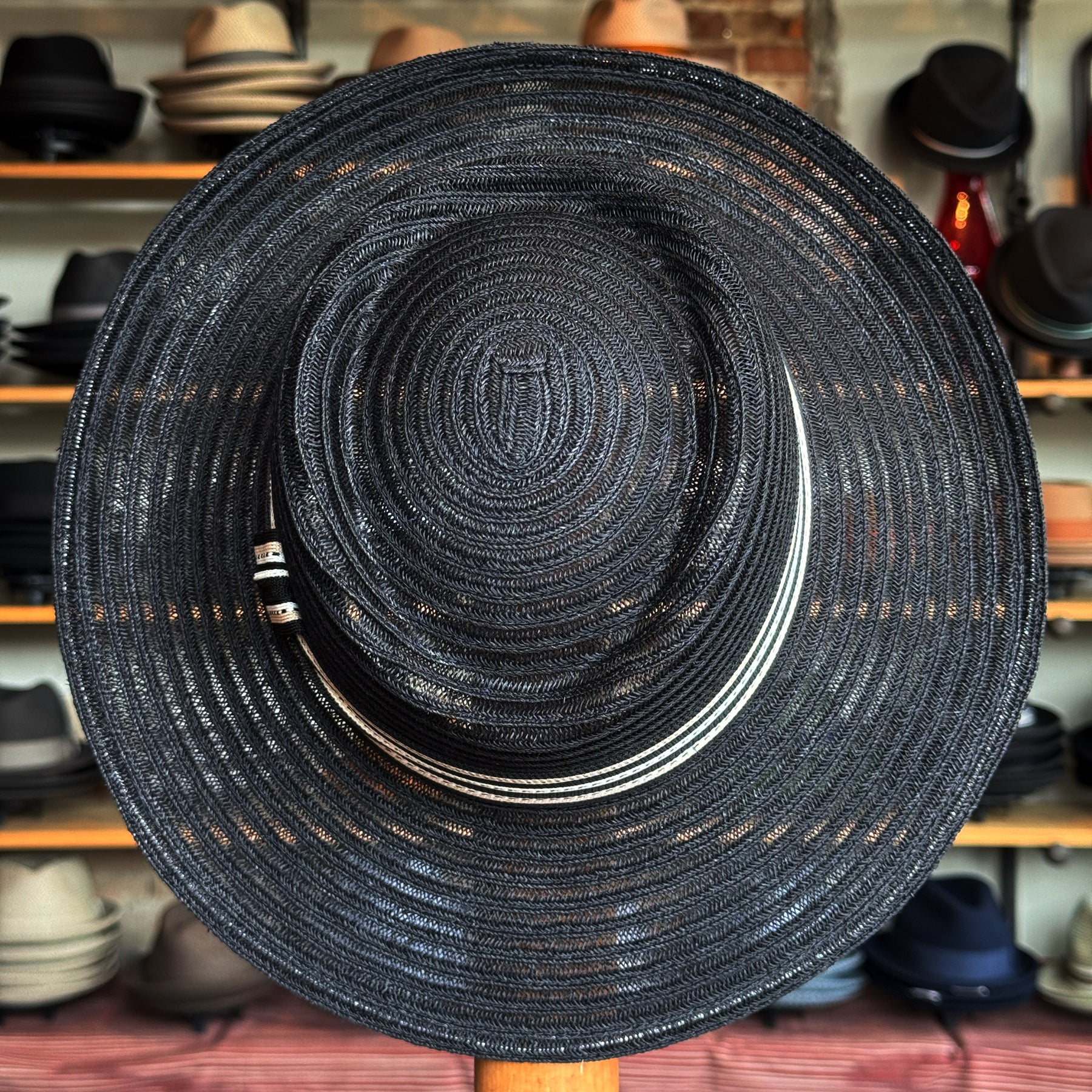Black woven Sabine Fedora with a wide brim and a decorative black and white band, displayed in a hat shop with various hats on shelves in the background.