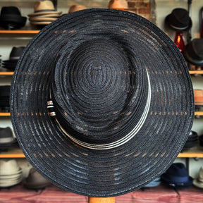Black woven Sabine Fedora with a wide brim and a decorative black and white band, displayed in a hat shop with various hats on shelves in the background.