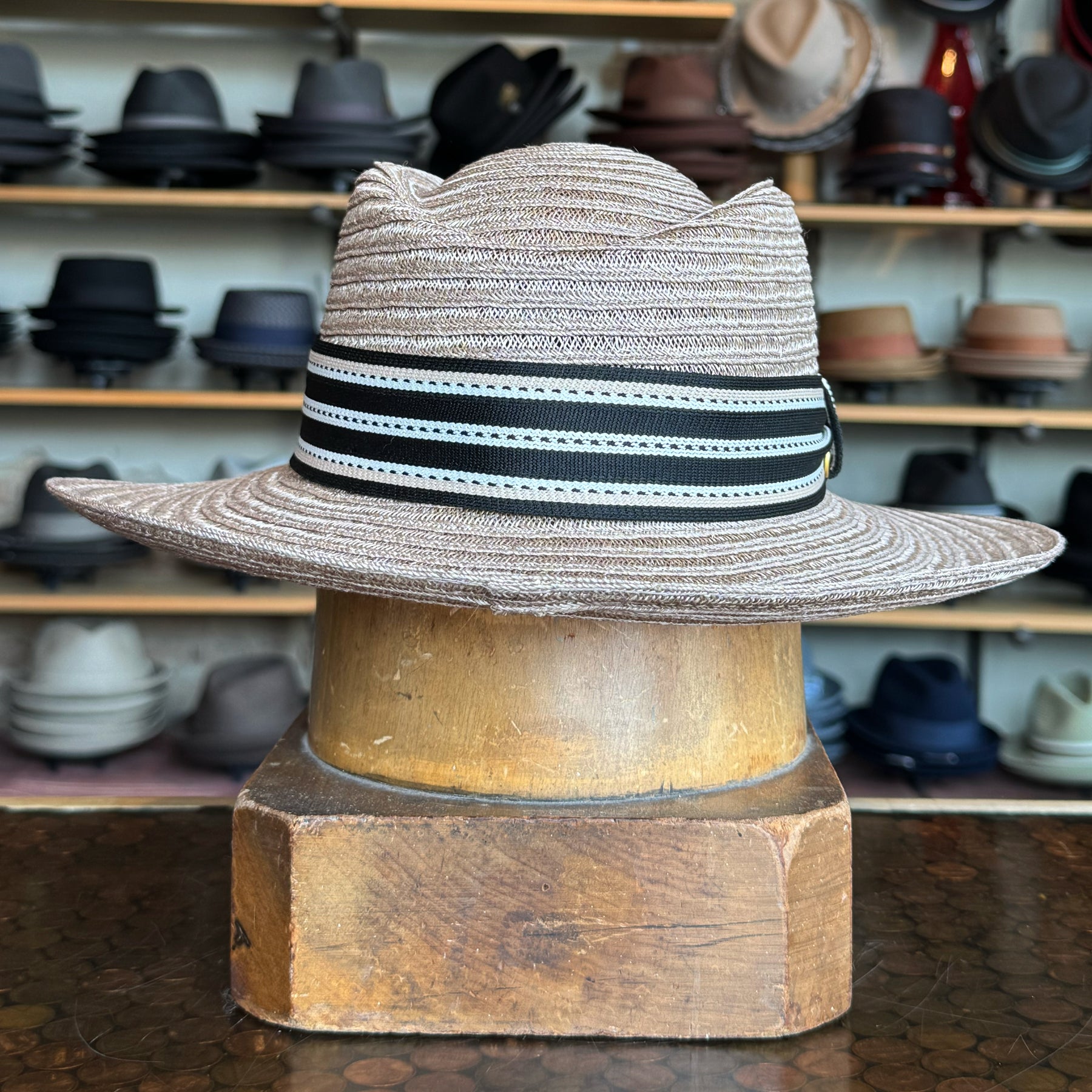 Sabine Fedora in taupe color with a black and white striped band, displayed on a wooden hat block with shelves of various hats in the background
