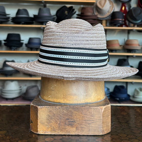 Sabine Fedora in taupe color with a black and white striped band, displayed on a wooden hat block with shelves of various hats in the background