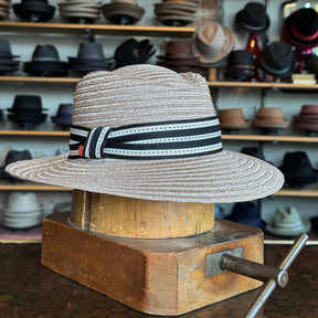 Sabine Fedora hat in taupe color with a black and white striped band, displayed on a wooden hat block in a shop with various hats in the background