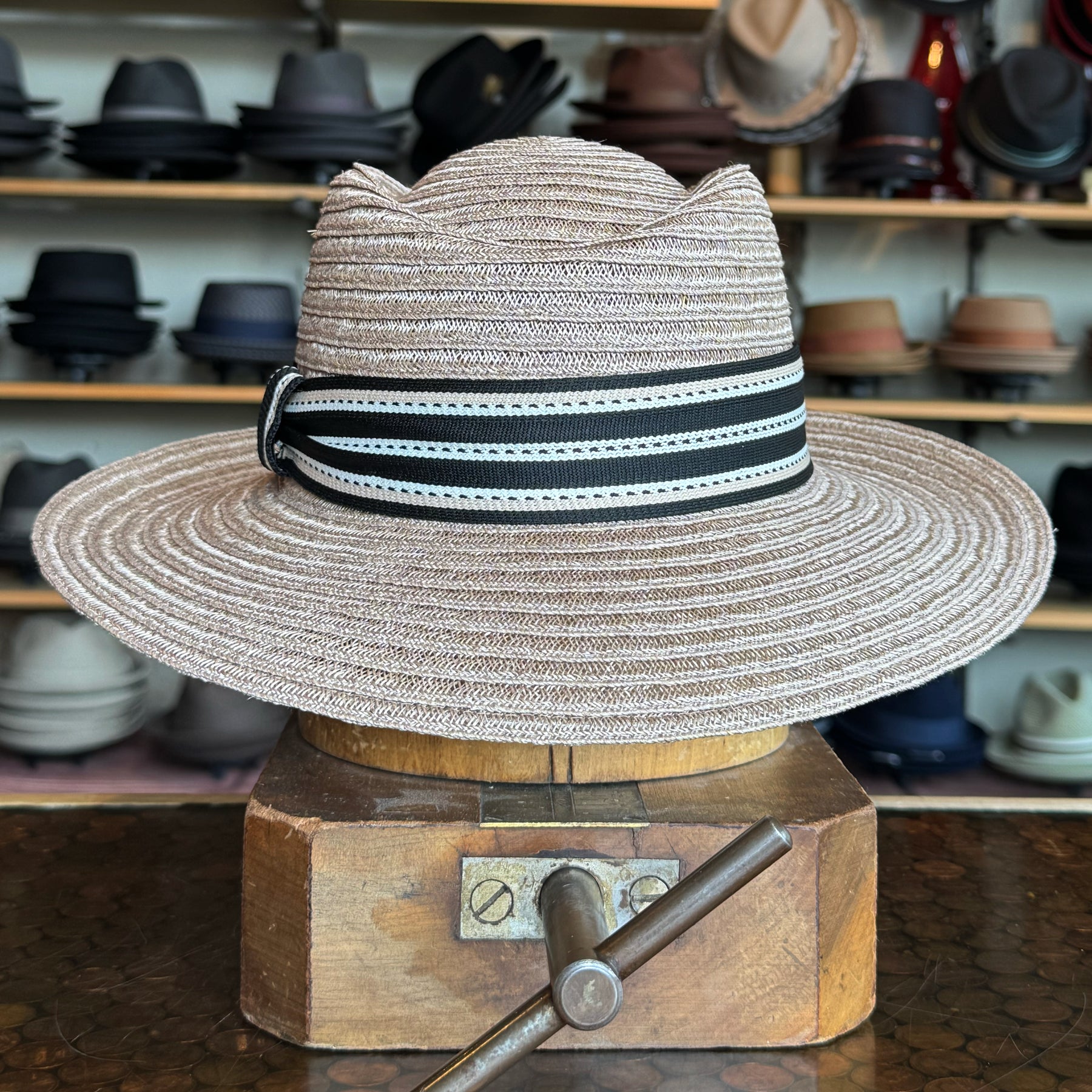 Sabine Fedora in taupe color with a wide brim and black-and-white striped band, displayed on a wooden hat block with various hats in the background.