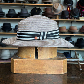 Sabine Fedora hat in taupe with a black and white striped band featuring a small red emblem, displayed on a wooden hat block in a shop filled with various hats.