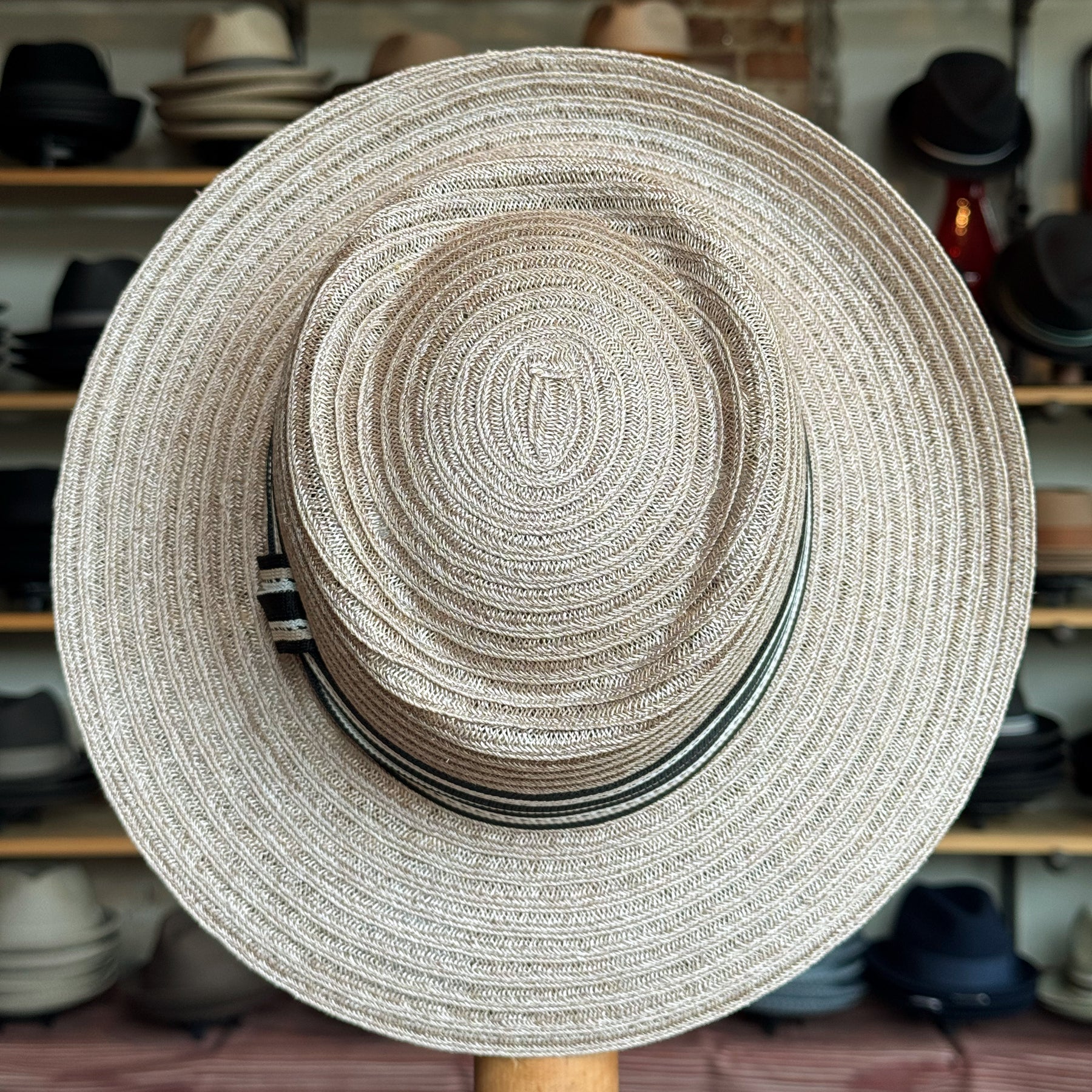 Sabine Fedora in taupe color featuring a wide woven brim and a subtle striped band, displayed in a hat shop setting with various hats in the background.