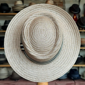 Sabine Fedora in taupe color featuring a wide woven brim and a subtle striped band, displayed in a hat shop setting with various hats in the background.