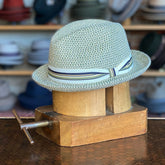 Salem woven straw hat with a light striped ribbon band, displayed on a wooden hat block in a shop with various hats in the background.