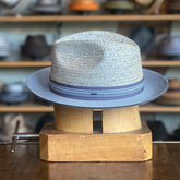 Scorsby Fedora in textured light grey with a subtle blue brim and striped ribbon band, displayed on a wooden hat block in a hat shop background.