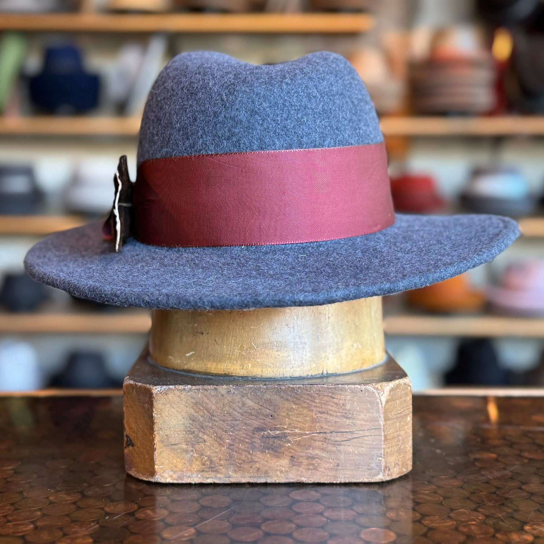 Sonora Fedora in gray felt featuring a wide burgundy band with a decorative bow, displayed on a wooden hat block with a softly blurred background of various hats.