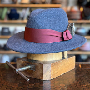 Sonora Fedora in charcoal gray felt with a wide brim and a burgundy ribbon band, displayed on a wooden hat block in a hat shop setting.