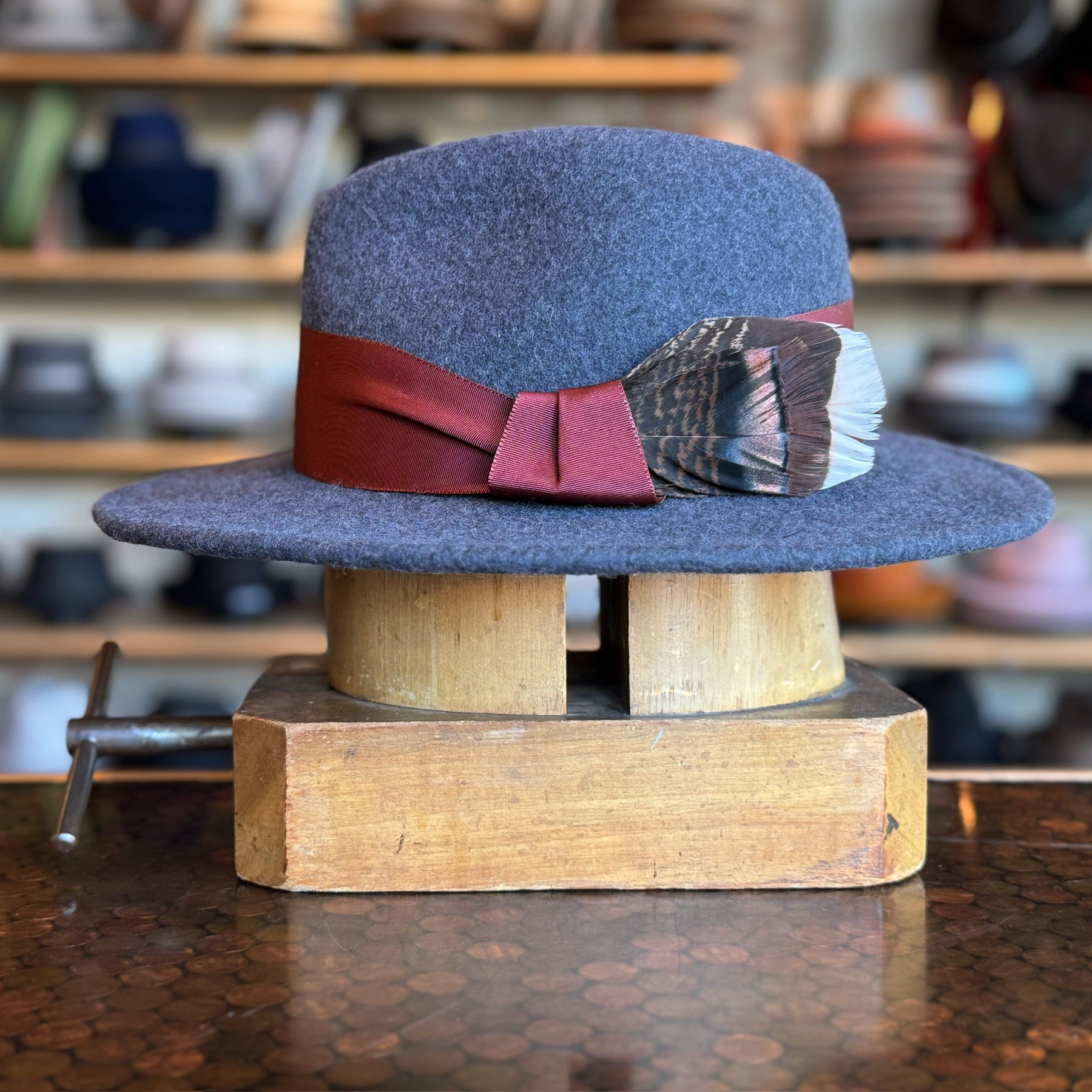 Sonora Fedora in charcoal gray felt with a wide brim, accented by a maroon band and decorative brown and white feather, displayed on a wooden hat block in a hat shop.
