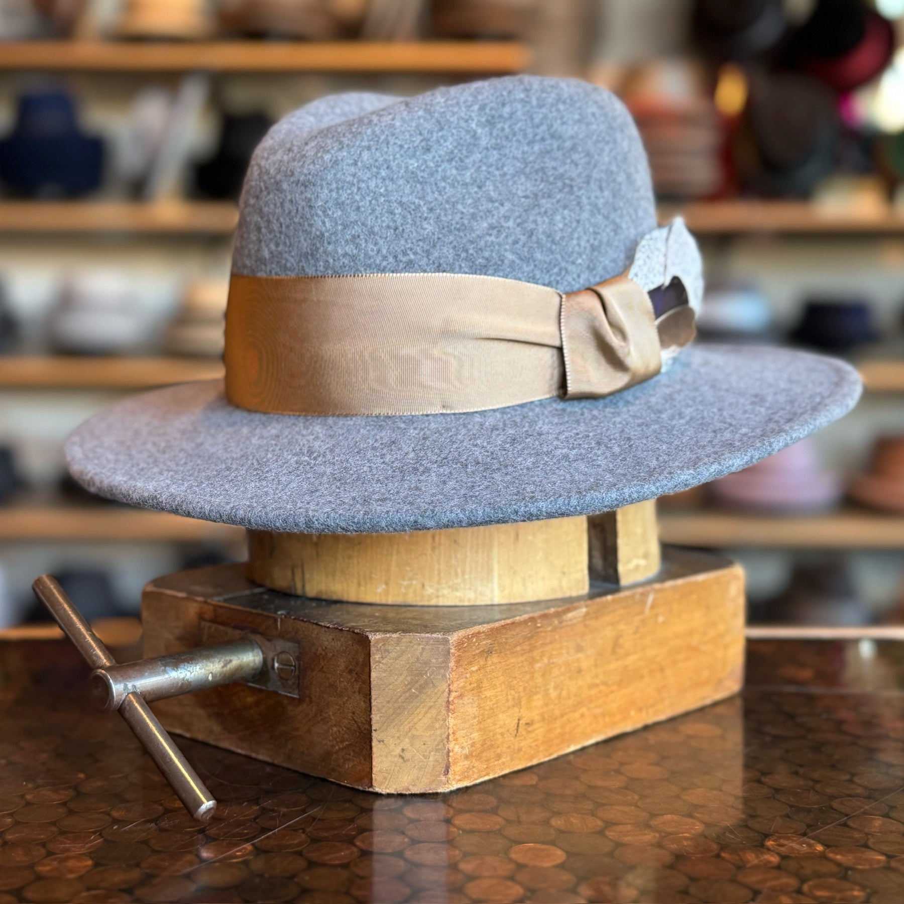 Sonora Fedora in soft grey felt with a wide beige ribbon band displayed on a wooden hat block in a cozy shop setting offering classic brim hat style.