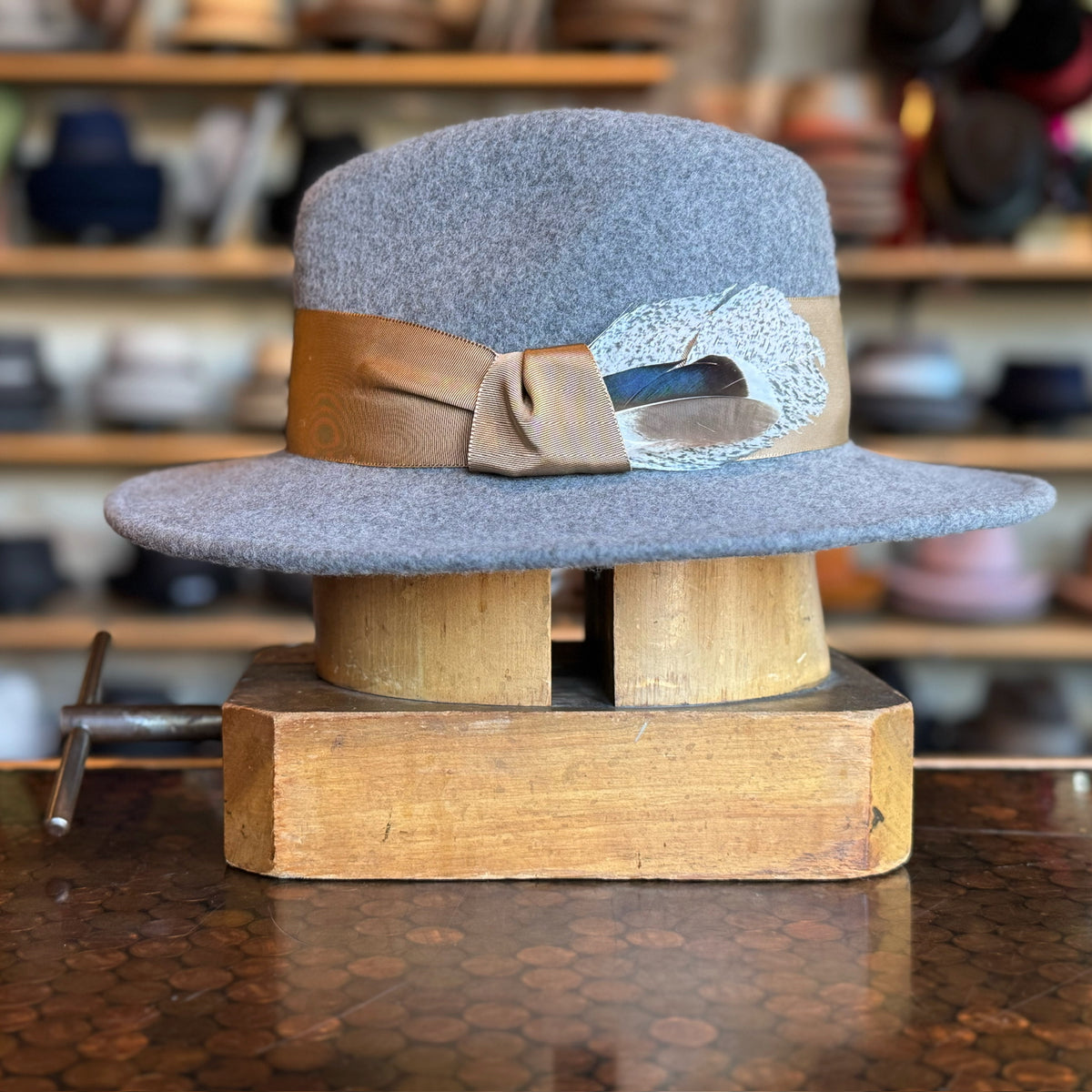 Sonora Fedora in grey felt with a brown ribbon band and decorative feather, displayed on a wooden hat block with shelves of hats in the background.