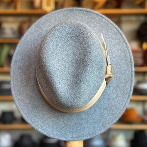 Light grey Sonora Fedora made of soft felt with a beige hatband and a feather detail, displayed against a blurred background of a hat shop interior.