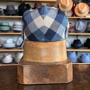 Squares Retro Flat Cap displayed on a wooden hat stand, featuring a blue and beige checkered pattern, in a background filled with various other hats and caps.