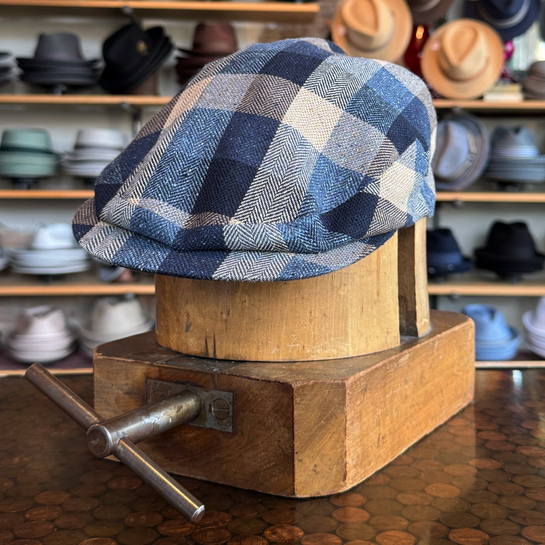 Squares Retro Flat Cap in a blue and white checkered pattern displayed on a wooden hat block with various hats blurred in the background, showcasing a stylish flat cap accessory.