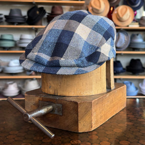 Squares Retro Flat Cap in a blue and white checkered pattern displayed on a wooden hat block with various hats blurred in the background, showcasing a stylish flat cap accessory.