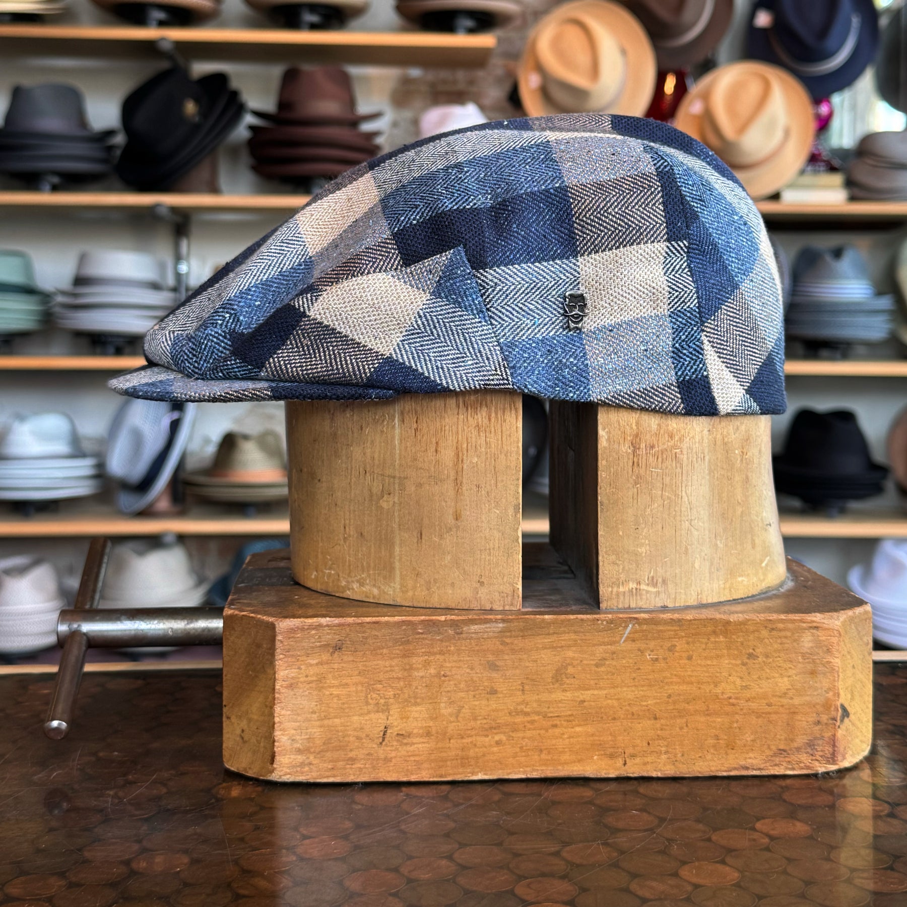 Squares Retro Flat Cap in blue and beige plaid pattern displayed on a wooden hat block, showcased in a store with various other hats in the background.