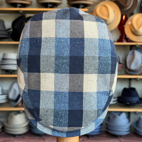 Squares Retro Flat Cap featuring a classic blue, cream, and brown checkered pattern, displayed on a wooden stand with various hats in the background.